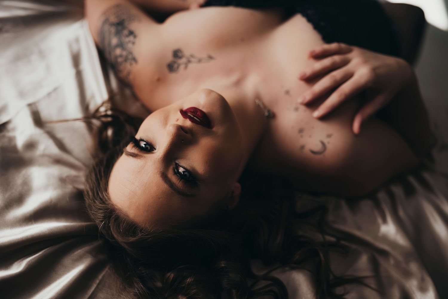 Close-up of a woman lying on satin sheets, her bold look highlighted by dark lipstick and intricate tattoos.