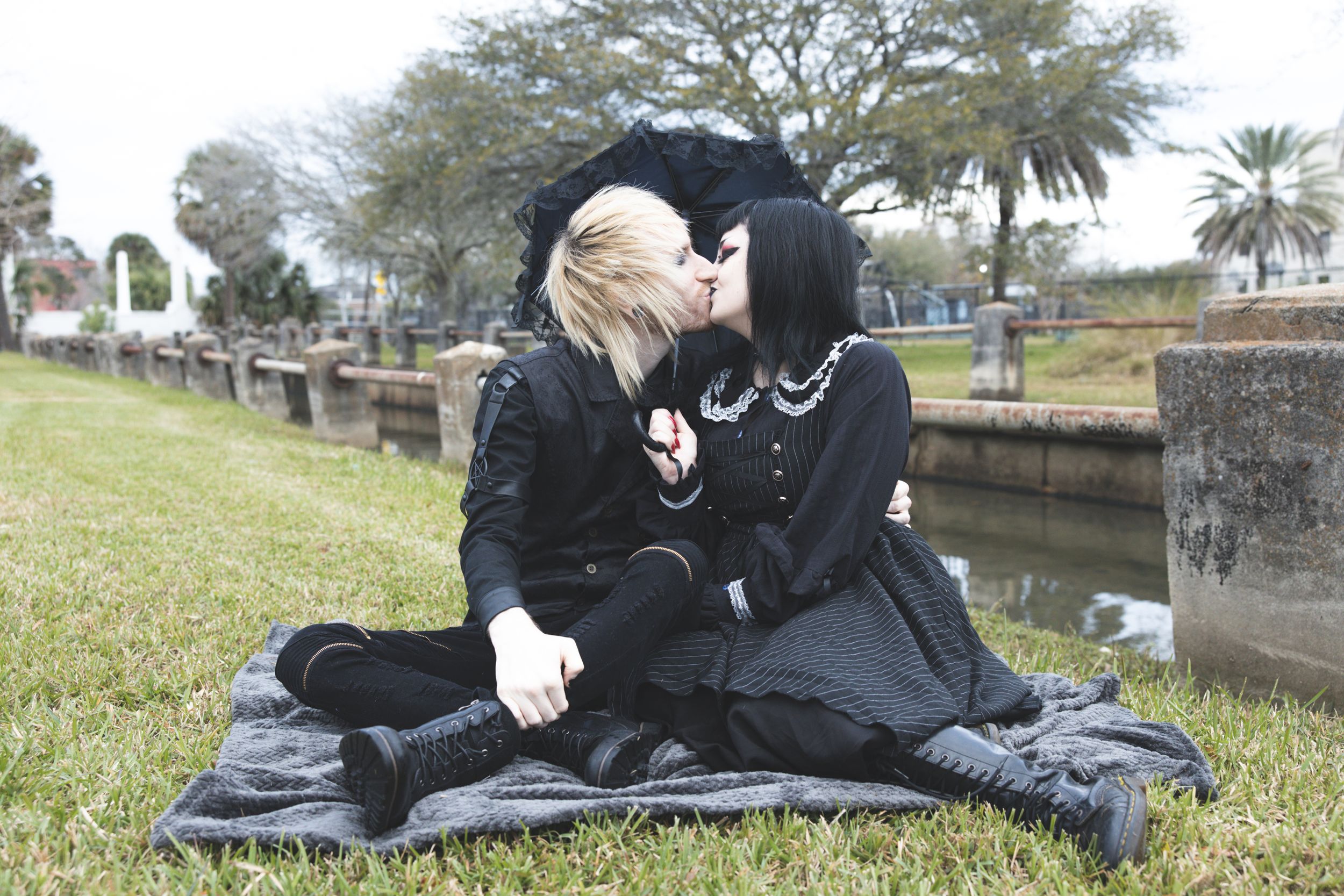 Two people in gothic attire share an intimate moment on a blanket by a canal.