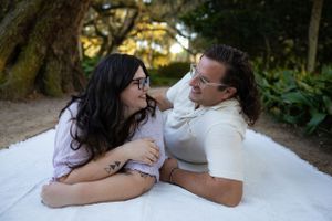 Couple laughing together while laying on white blanket in park setting.