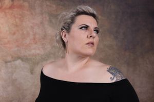 Dramatic portrait against textured brown wall featuring short blonde hair and shoulder tattoo.