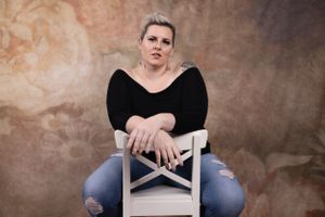 Artistic studio portrait showing contemplative pose on white chair against brown backdrop.