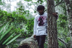 Person in white graphic hoodie poses against tree in lush forest setting.