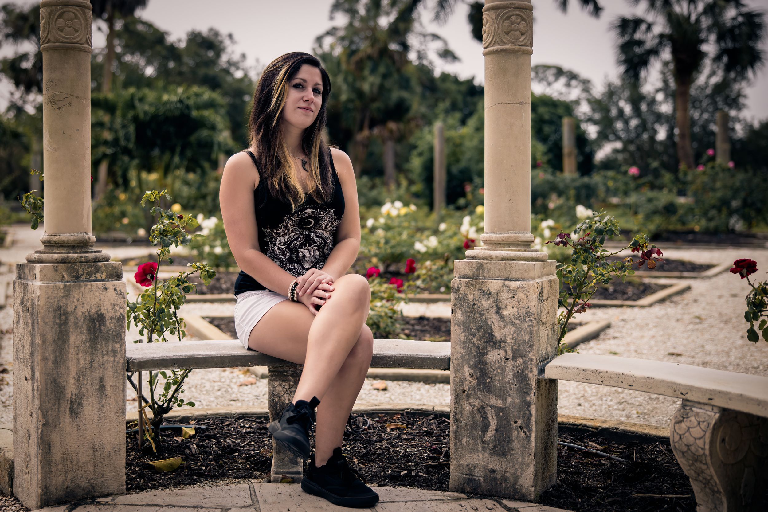 Young woman in dark dress sitting on stone bench in ornate garden with classical columns.