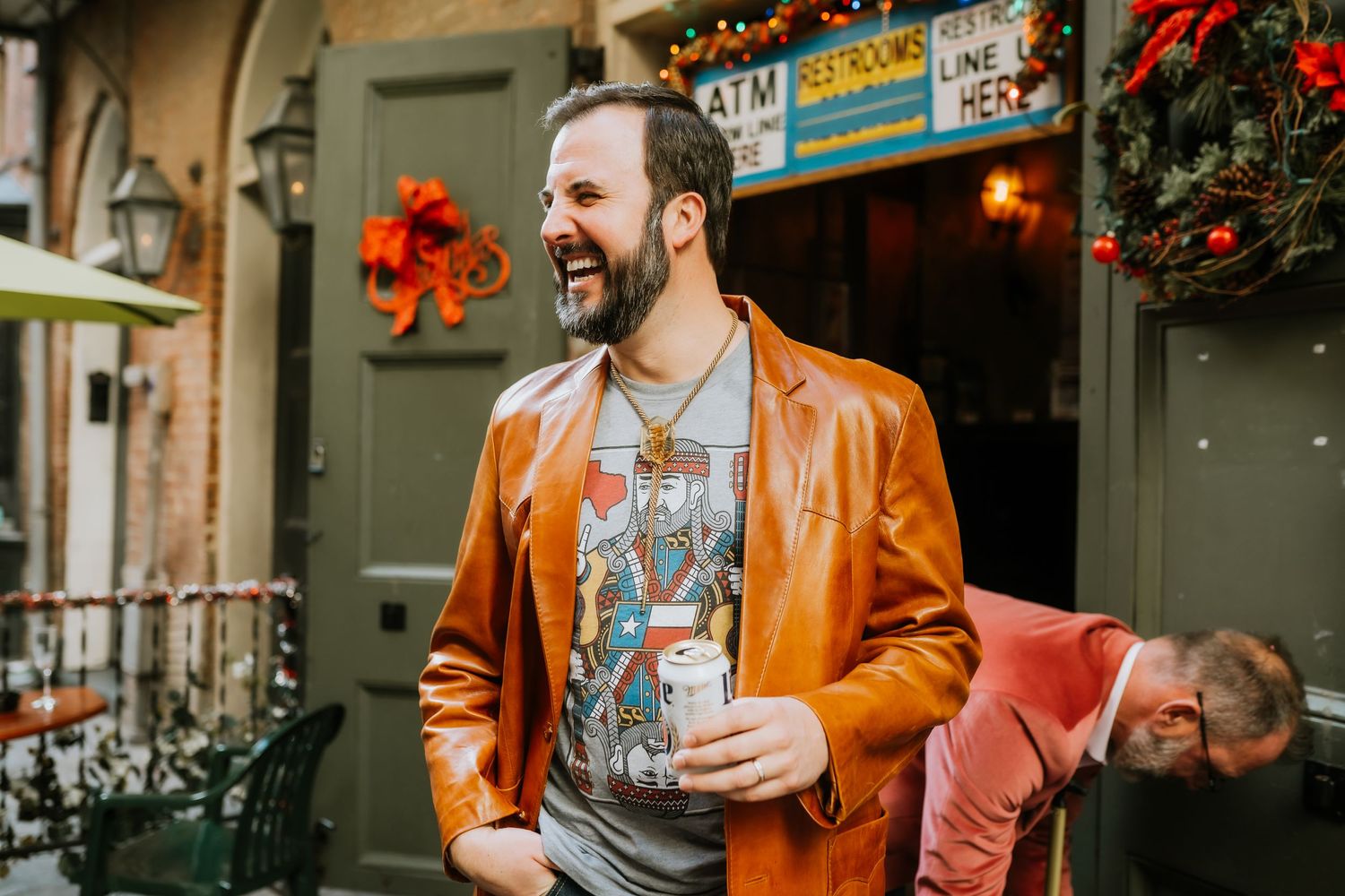 Person in bright orange leather jacket smiling while holding a drink outside a festive storefront.