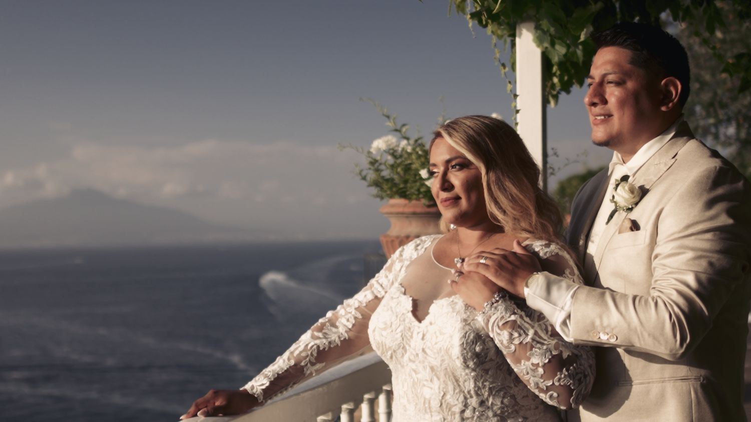 Bride and groom share romantic moment on balcony overlooking coastal landscape.