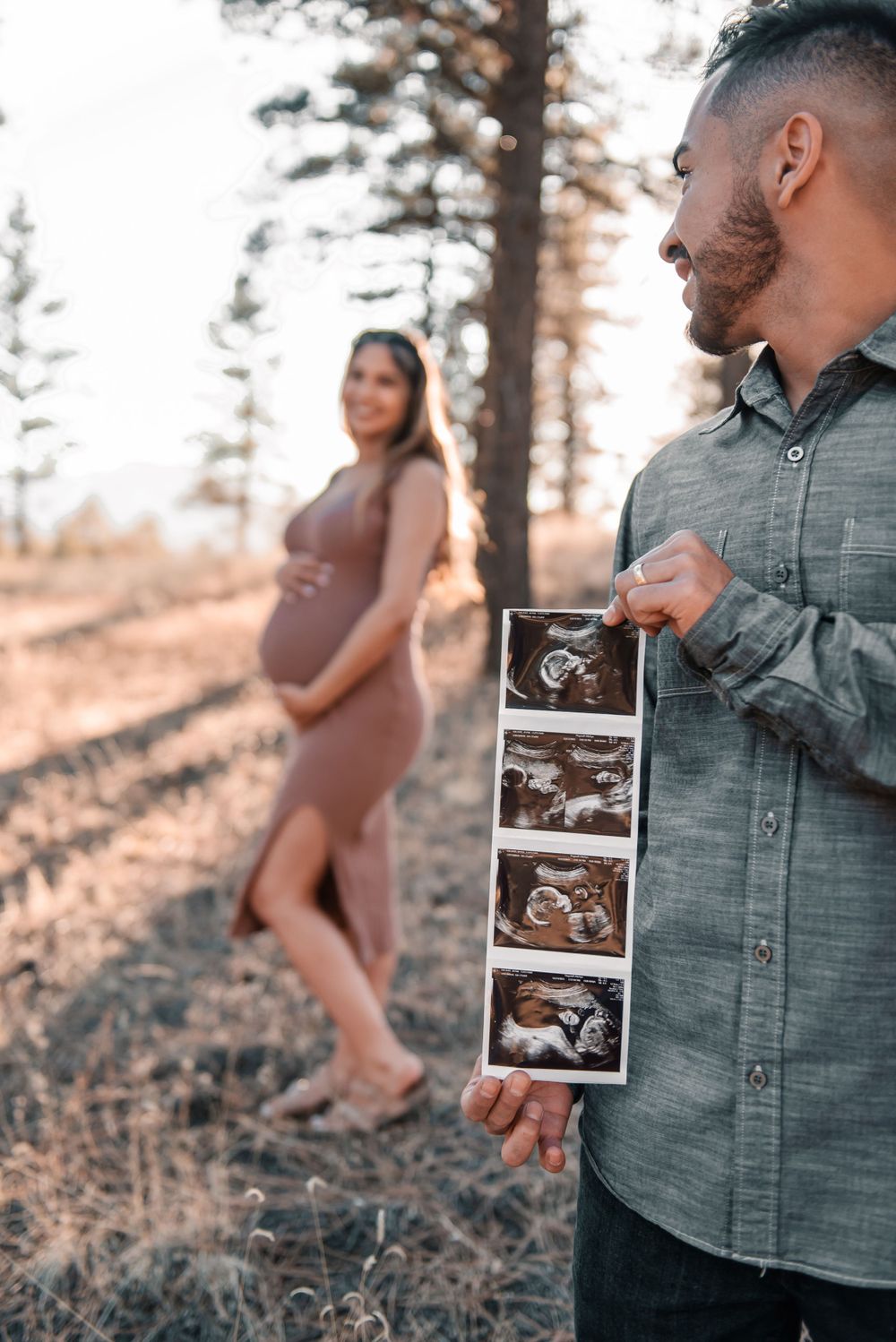 Couple holding ultrasound photos during outdoor maternity session among pine trees.