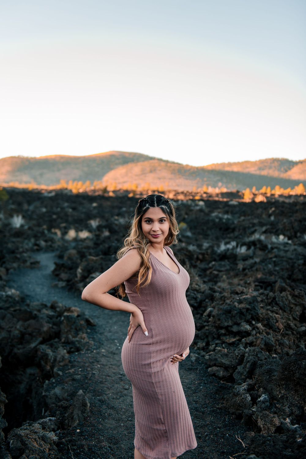 Expectant mother in pink dress poses against dramatic mountain landscape at dusk.