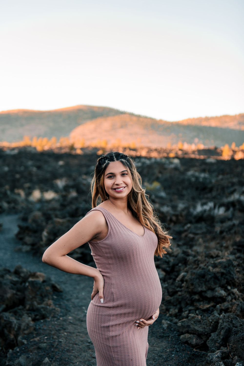 Maternity portrait on dark volcanic terrain with sunlit mountains in background.