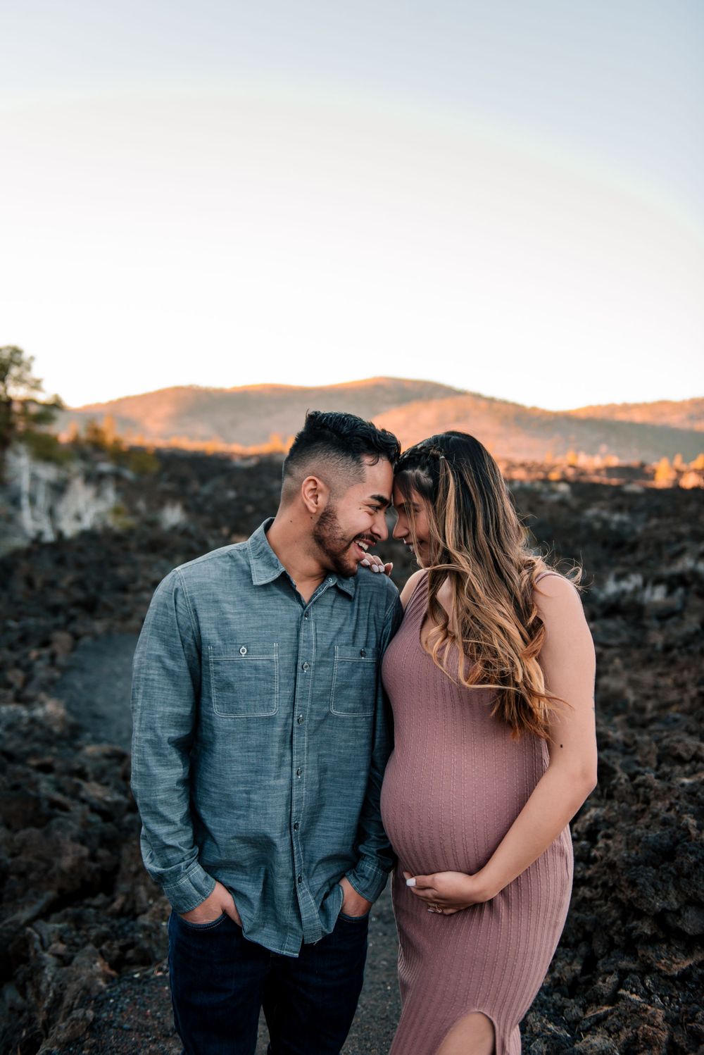 Couple shares intimate moment during sunset maternity shoot on volcanic rocks.