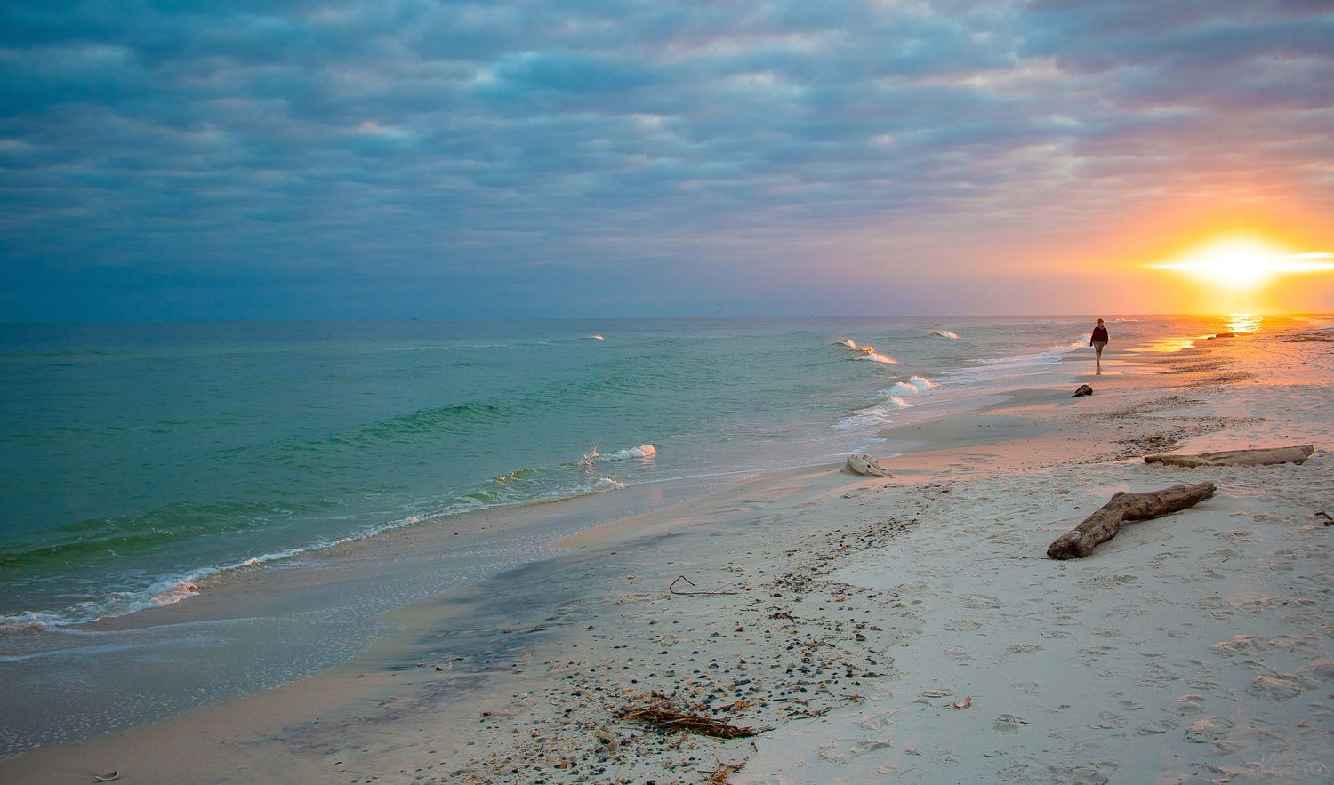 Tranquil beach sunset with gentle waves and a lone figure walking along the shoreline.