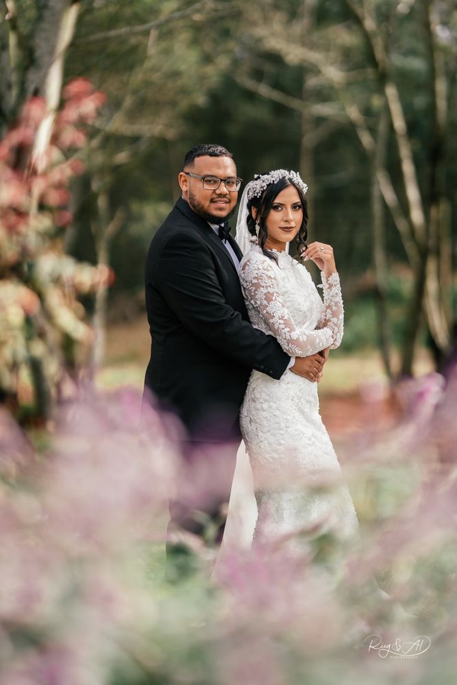 Elegant couple posing in a garden with purple flowers, groom in tuxedo and bride in lace wedding gown.