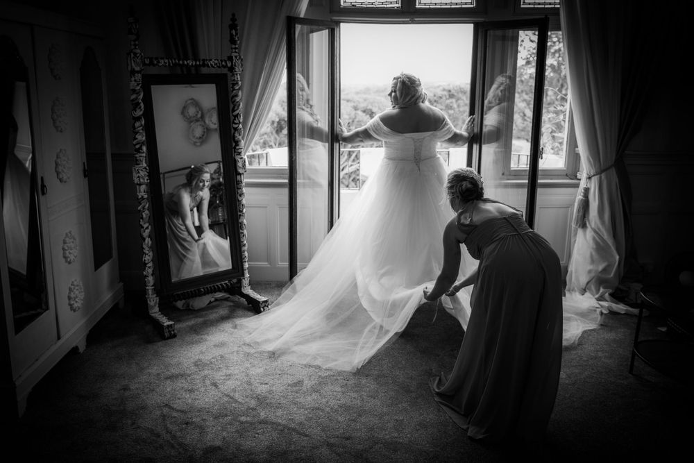 Black and white image showing wedding dress preparation near bright window.