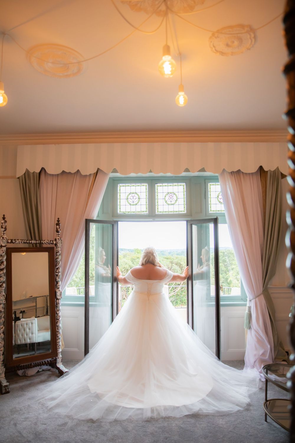 Wedding dress silhouette against bright window with flowing train and elegant curtains.