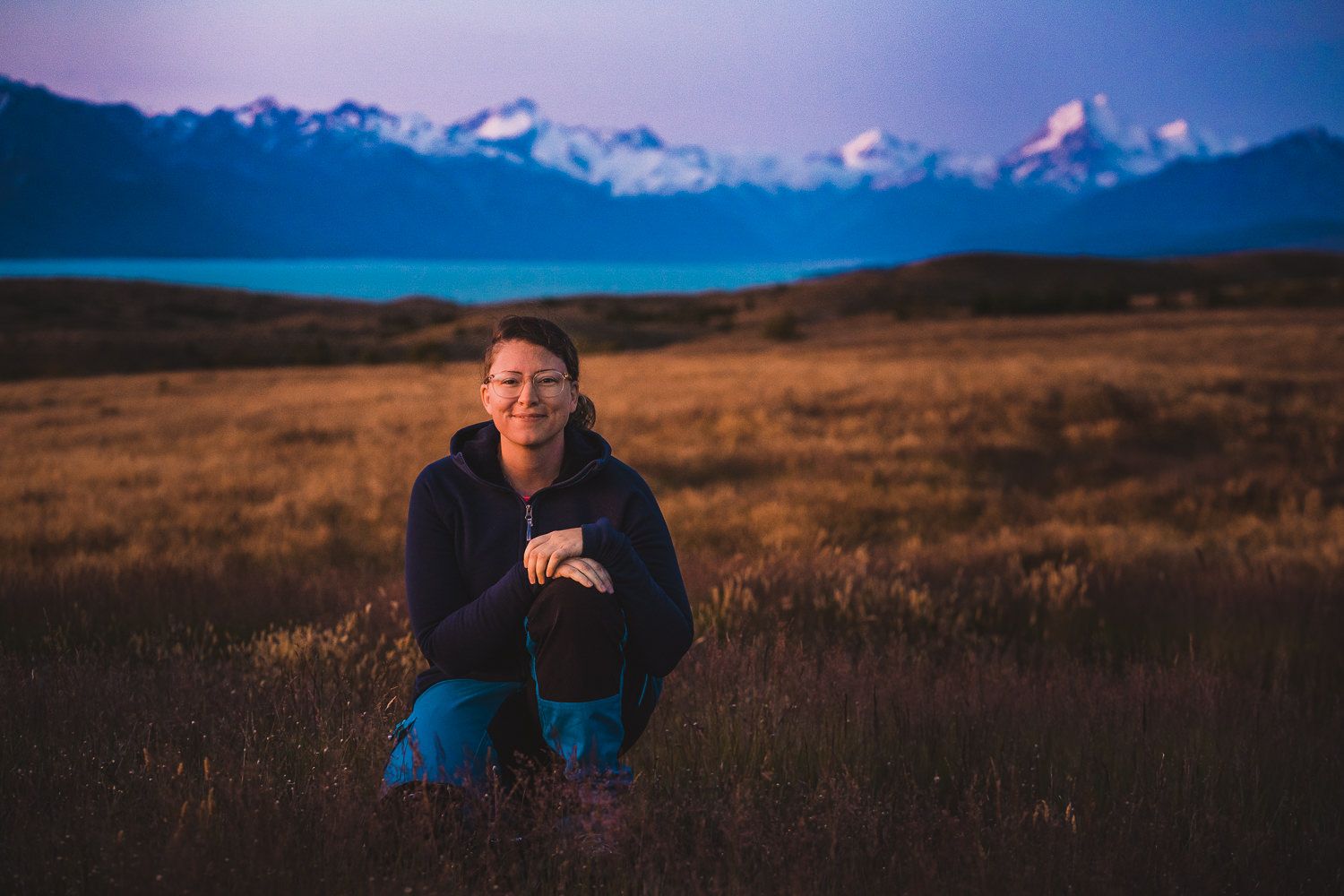 Fotograf Maria Karlsson sitter på huk framför Mount Cook , Nya Zeeland med rosa solnedgångshimmel och turkosblått vatten