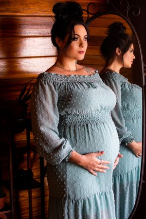 A maternity photoshoot in front of a wood wall featuring a light blue dotted dress with smocked details and mirror reflection.
