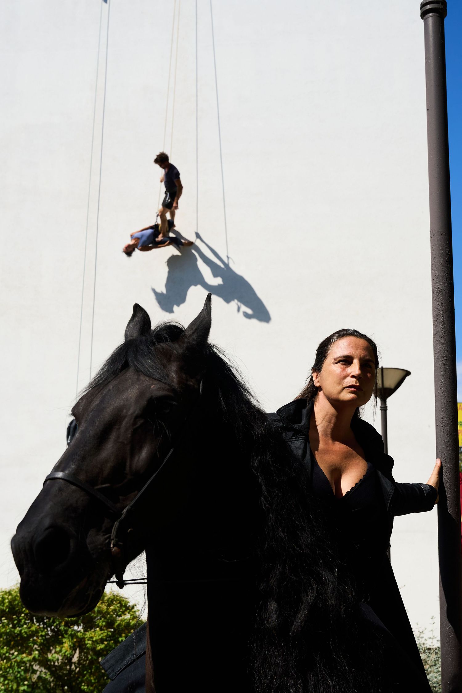 Dramatic scene with black horse and performer in costume against stark white background with suspended figure.