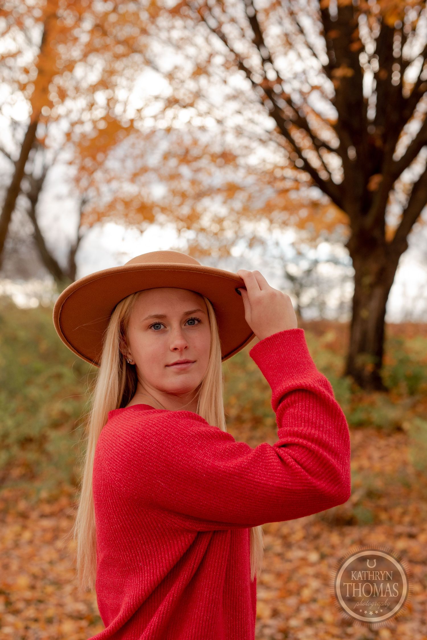 Portrait of someone in a red sweater and brown hat posing against fall foliage in a natural outdoor setting.