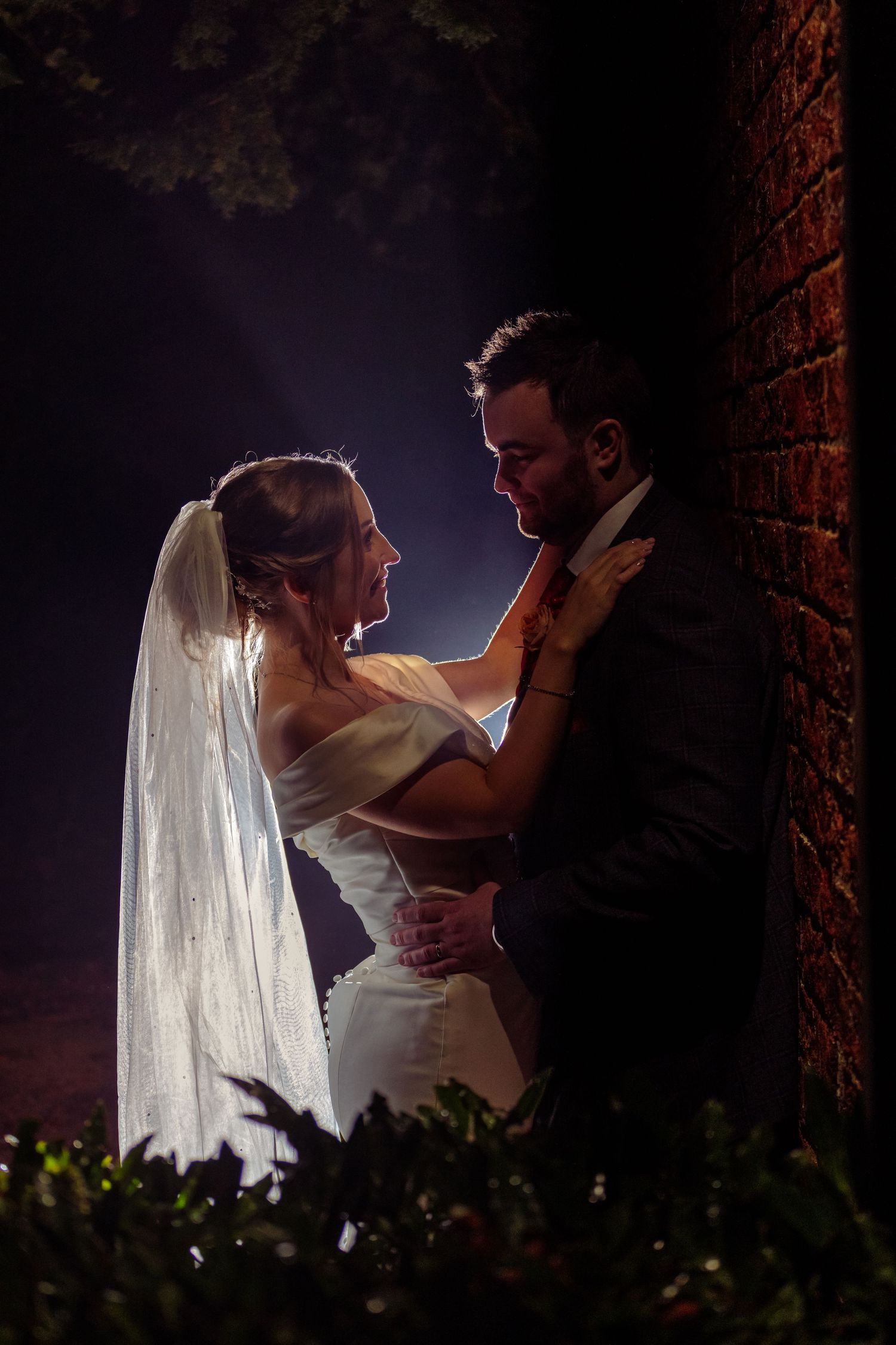 A romantic backlit silhouette of newlyweds sharing an intimate moment against a brick wall backdrop.