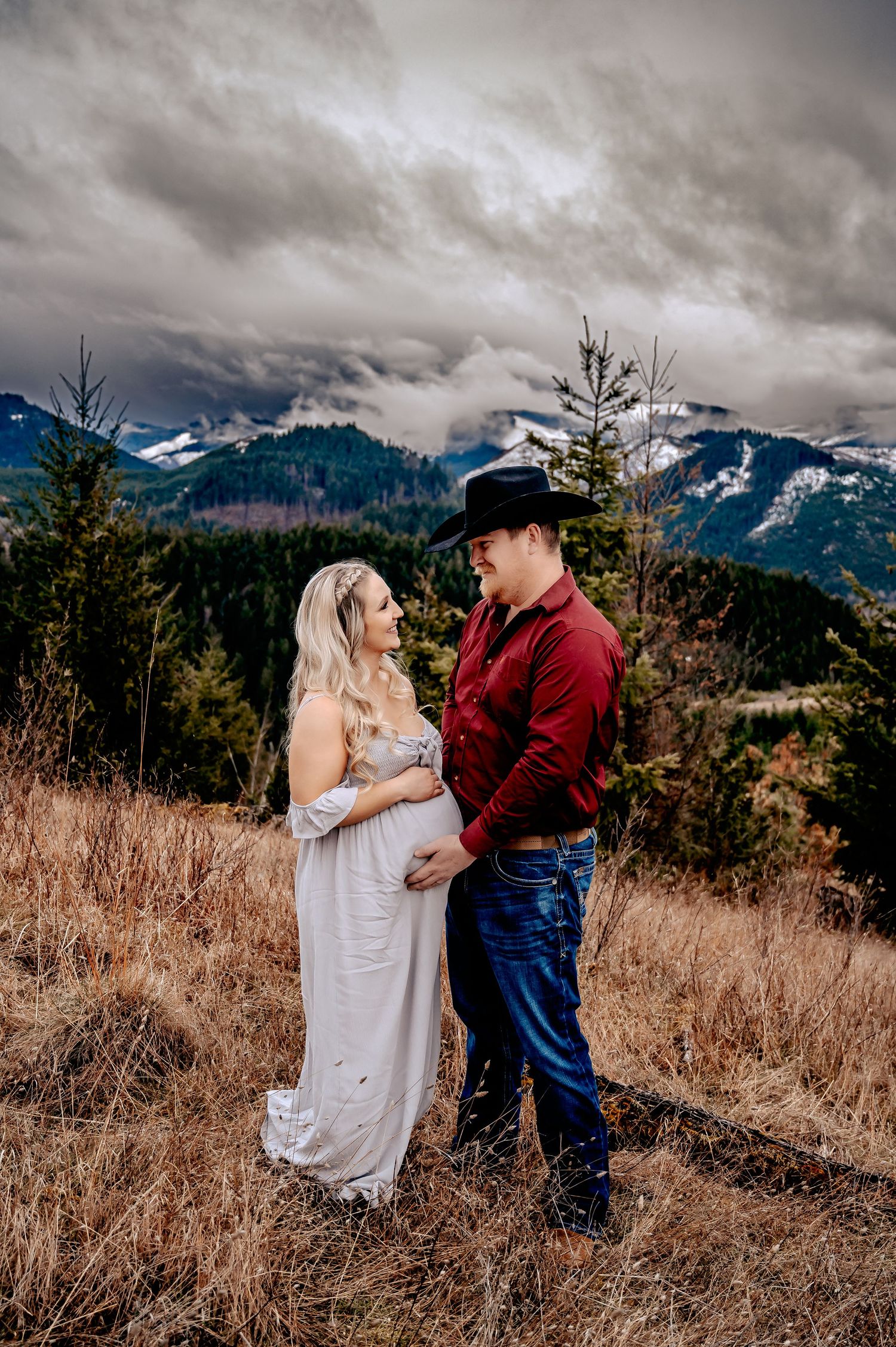 Couple standing in golden field with snow-capped mountains and dramatic clouds in background.
