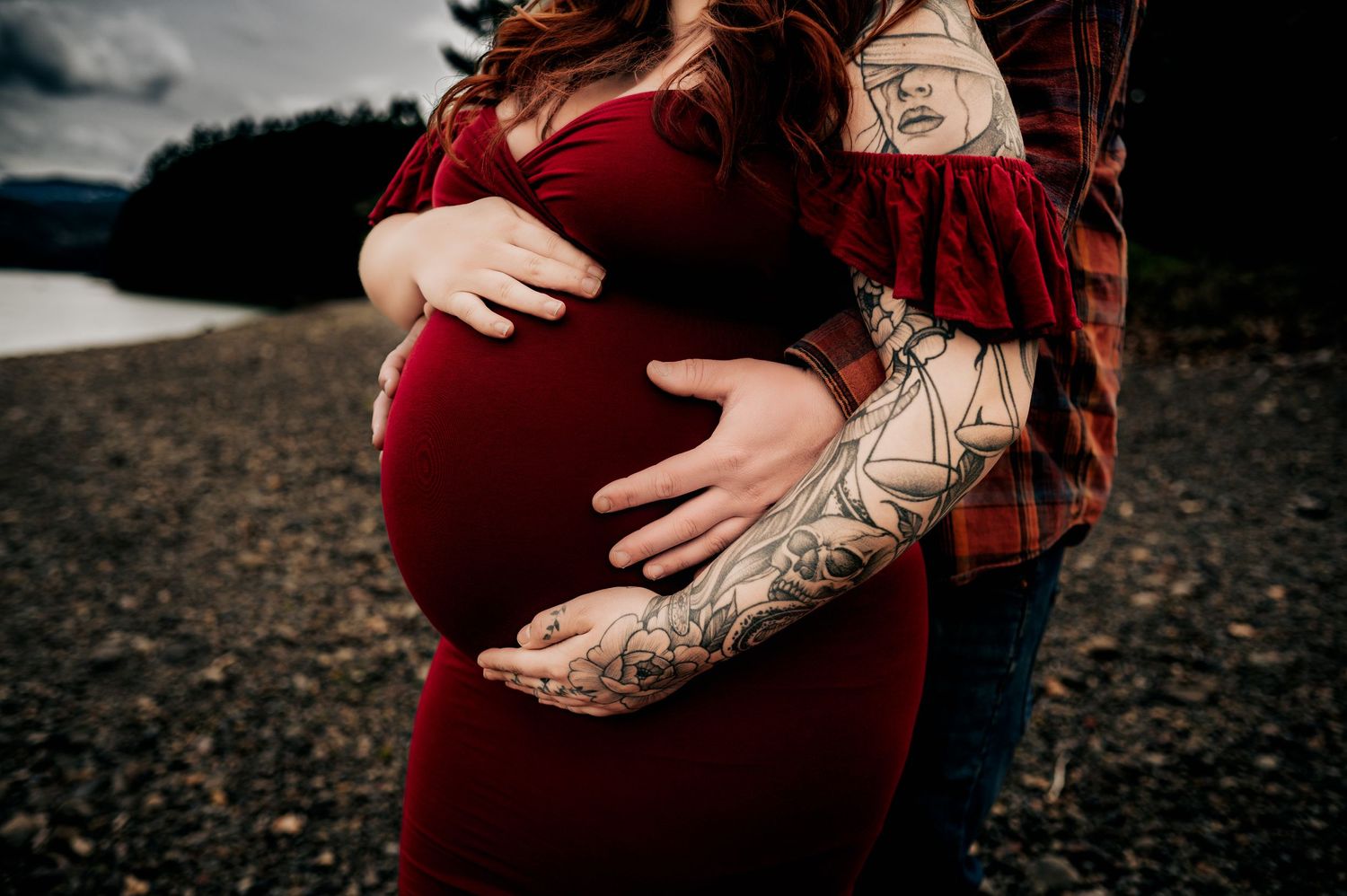 Close-up of tattooed arms cradling a pregnant belly against dark background.