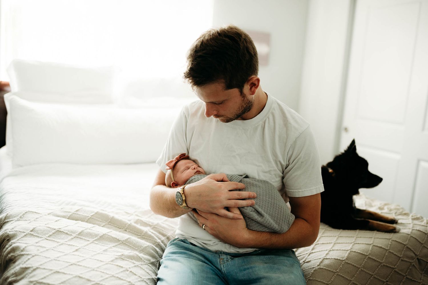 Parent in white shirt holding sleeping infant on white bedding in natural light.