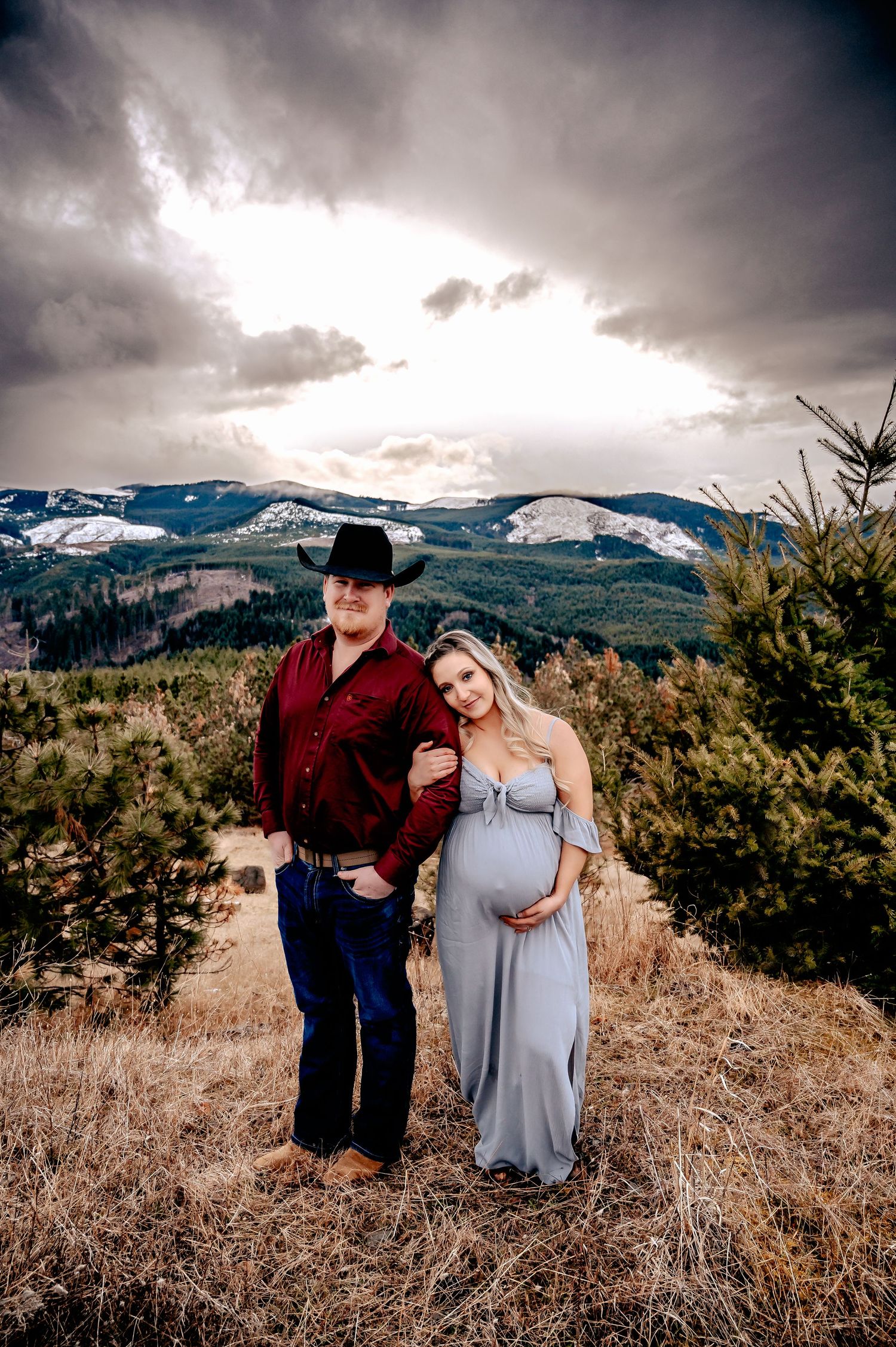 A couple poses together on a hilltop with snow-capped mountains and dramatic clouds in the background.