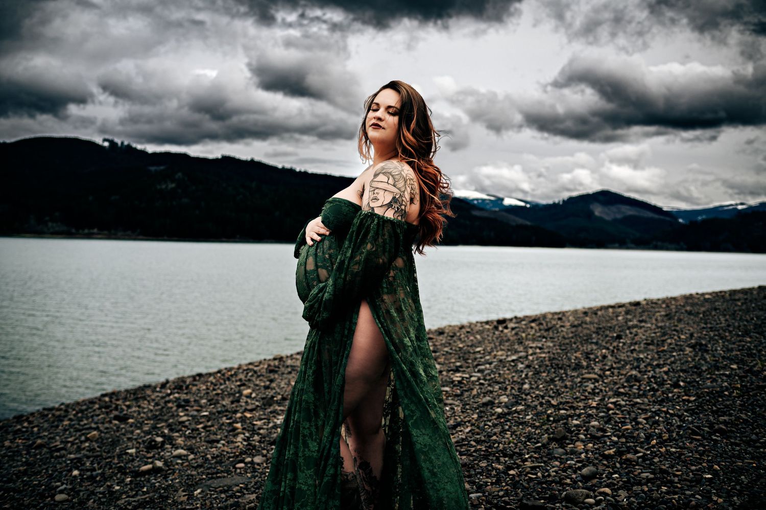 Person in emerald green dress walks along a rocky shoreline under moody storm clouds.