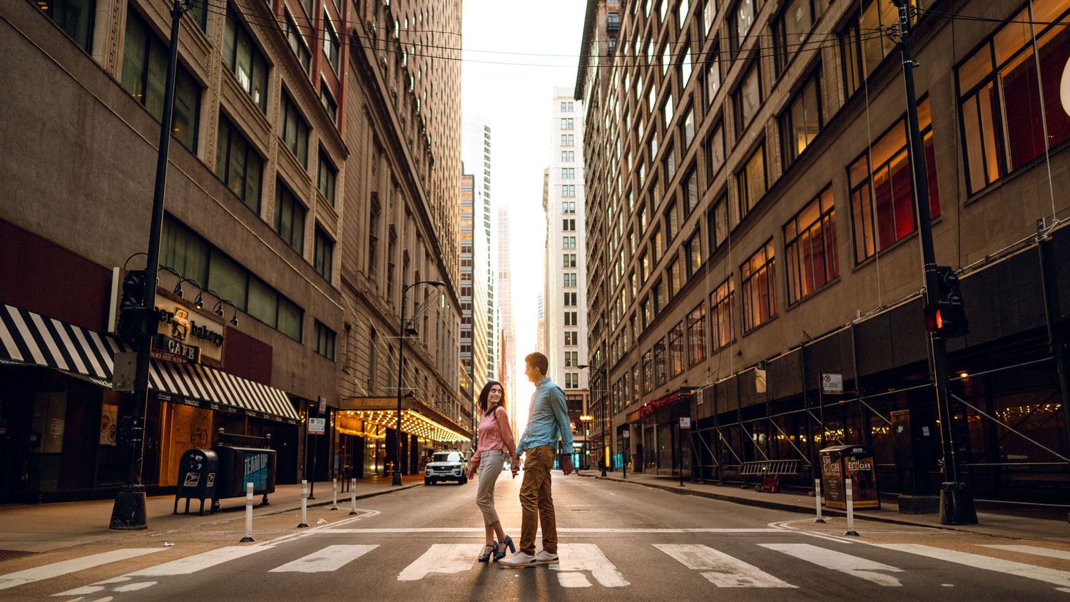 Couple strolls Chicago streets, lady's glances back at man as the sun's warm glow dances through the tunnel of buildings