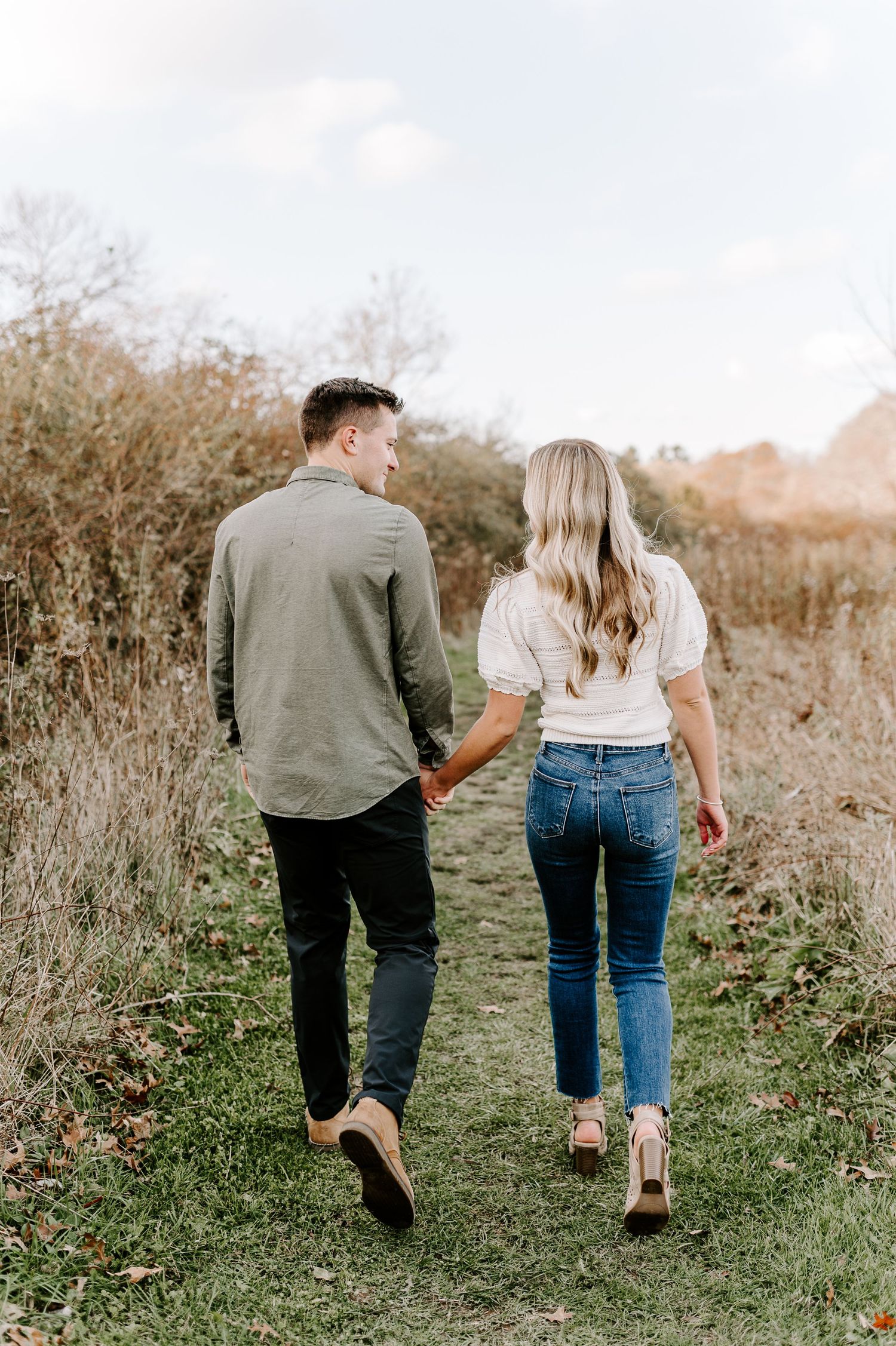 Couple walking hand in hand along a grassy trail during an outdoor photo session.