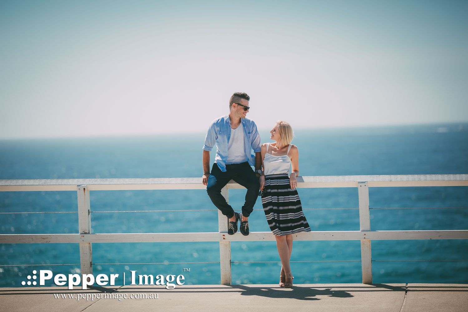Couple sitting on white railing by the ocean on a bright sunny day at the waterfront.