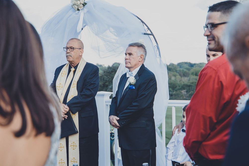 Wedding ceremony taking place under an outdoor arch with guests seated on white chairs.