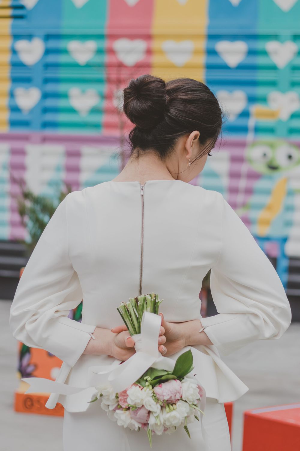 Two people in white outfits pose with floral bouquets against a vibrant mural with heart patterns.