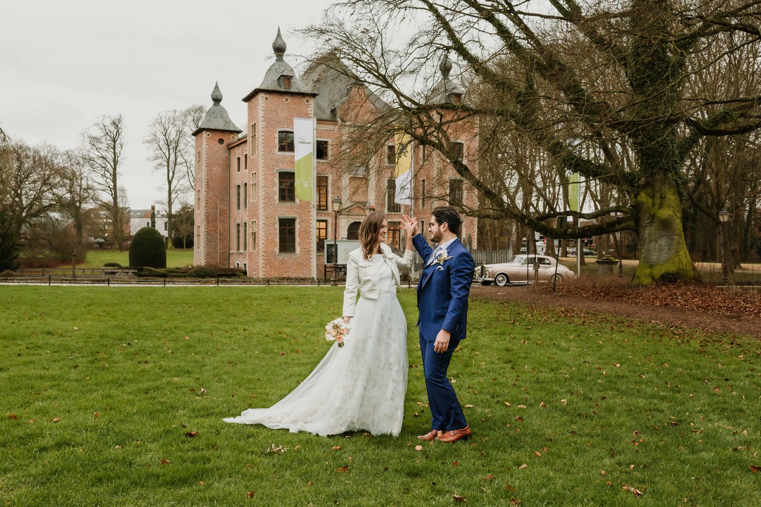 A wedding couple poses in front of a stately pink castle on a lush green lawn.