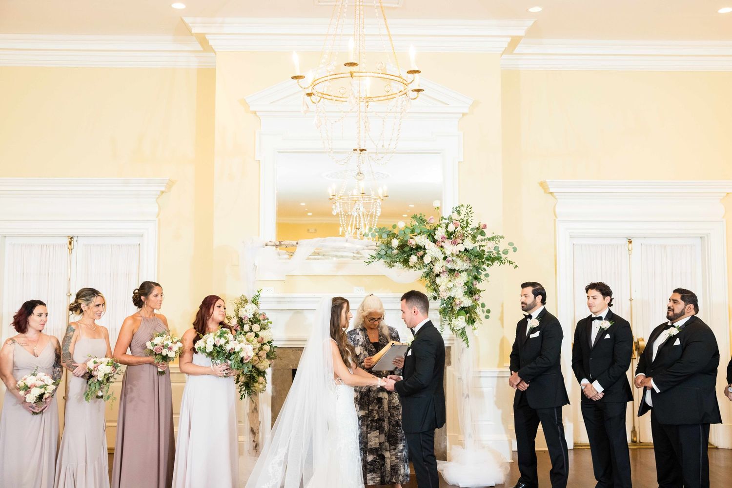 Indoor wedding ceremony in elegant yellow room with chandelier and floral arrangements.
