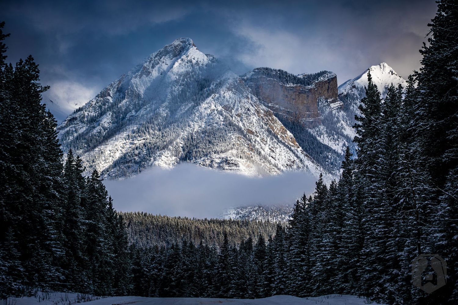 Snow-capped mountain peak towers over a winter forest landscape with dramatic cloudy skies in the background.
