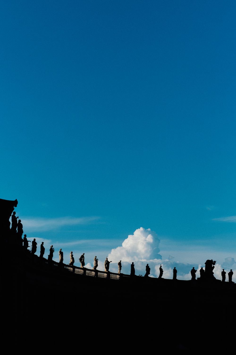 Silhouettes of statues line up against bright blue sky atop a historic Roman building.