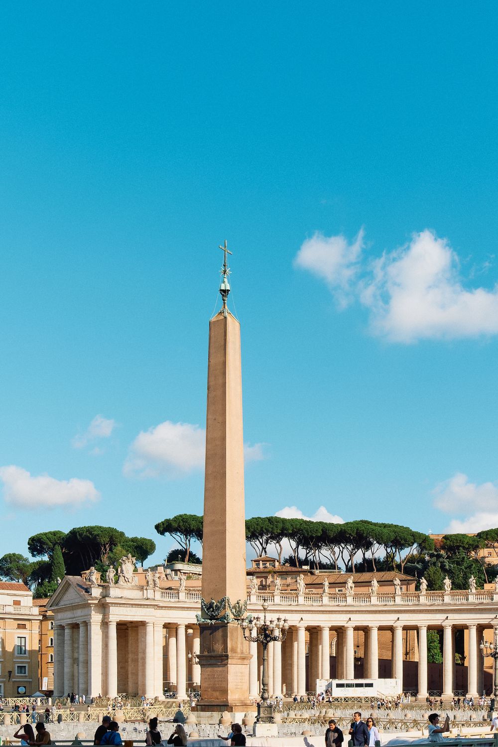 Vatican obelisk stands tall against blue sky with Saint Peter's Square colonnade and basilica in background.