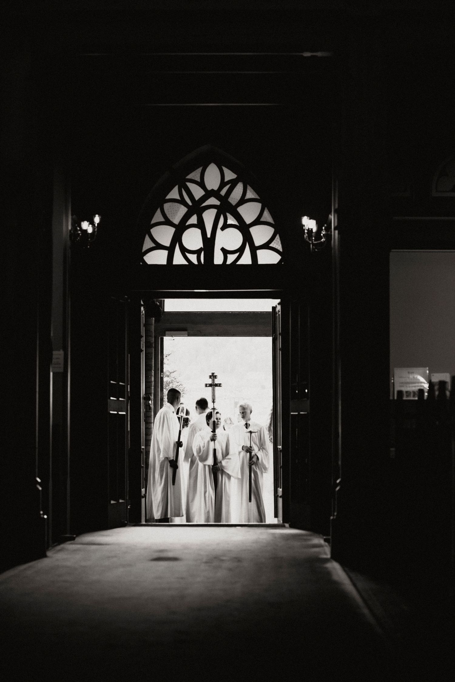 Dramatic silhouette of robed figures in a dark church corridor with gothic arched windows.
