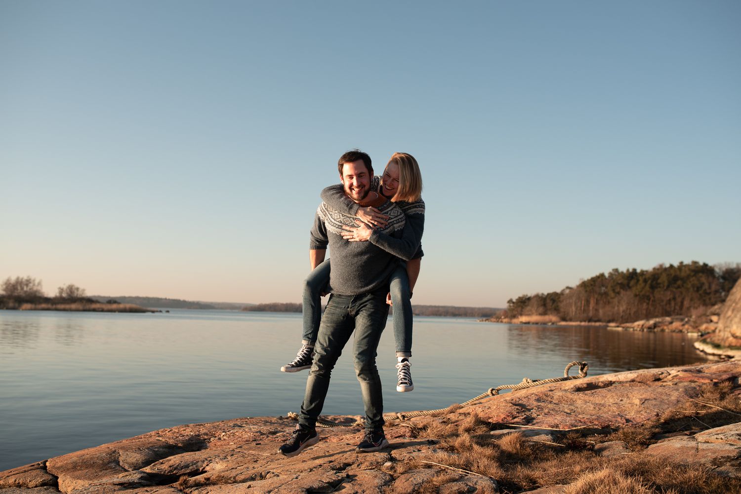 Joyful couple sharing playful moment on rocky waterfront during sunset.