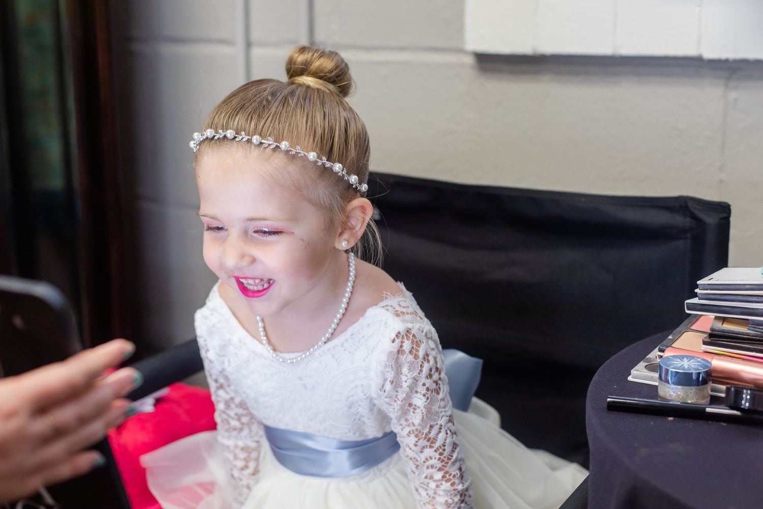 A joyful flower girl wearing a white lace dress with a silver sash and pearl headpiece smiles during wedding preparations.