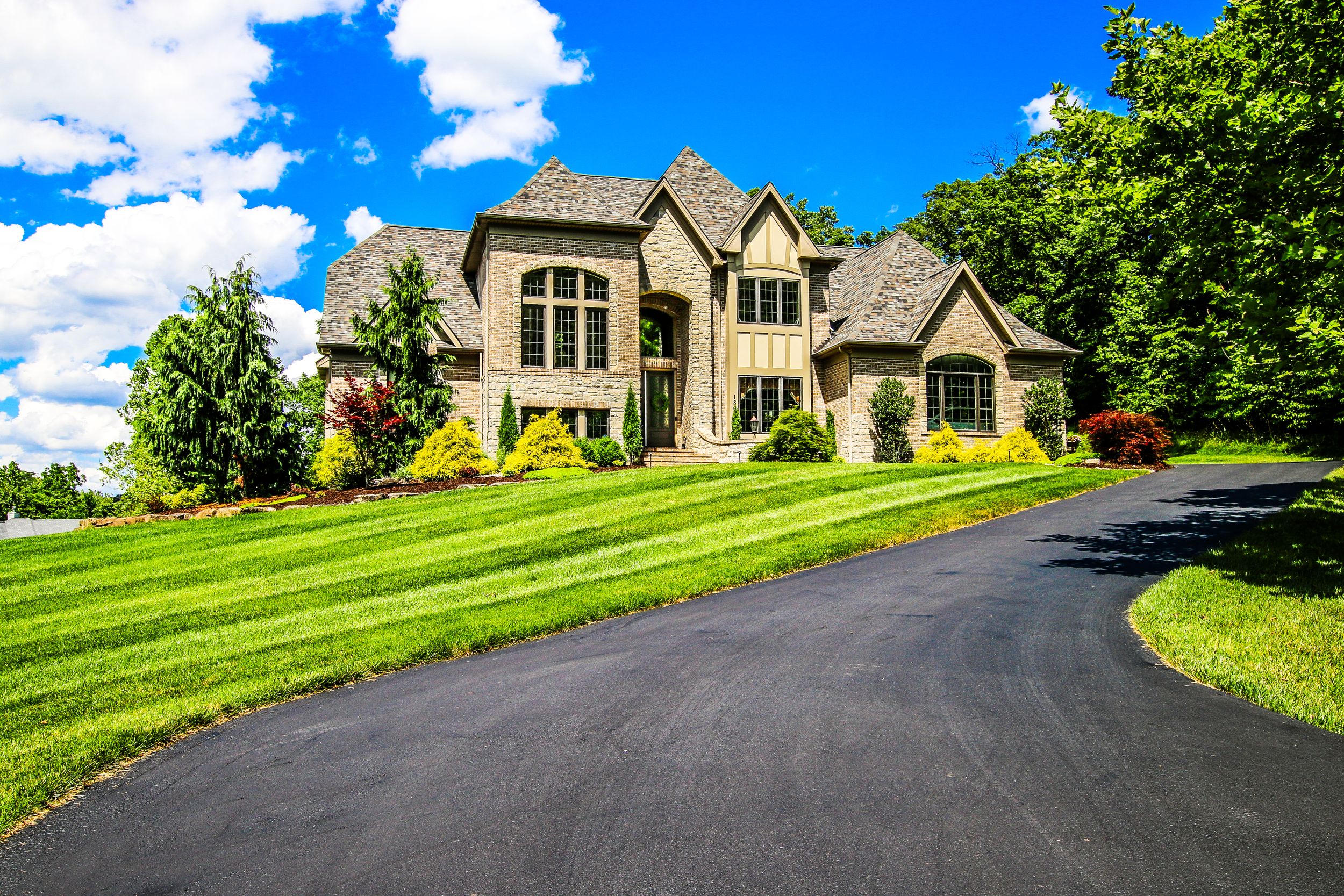 Luxury stone mansion with manicured lawn and curved driveway on sunny day.
