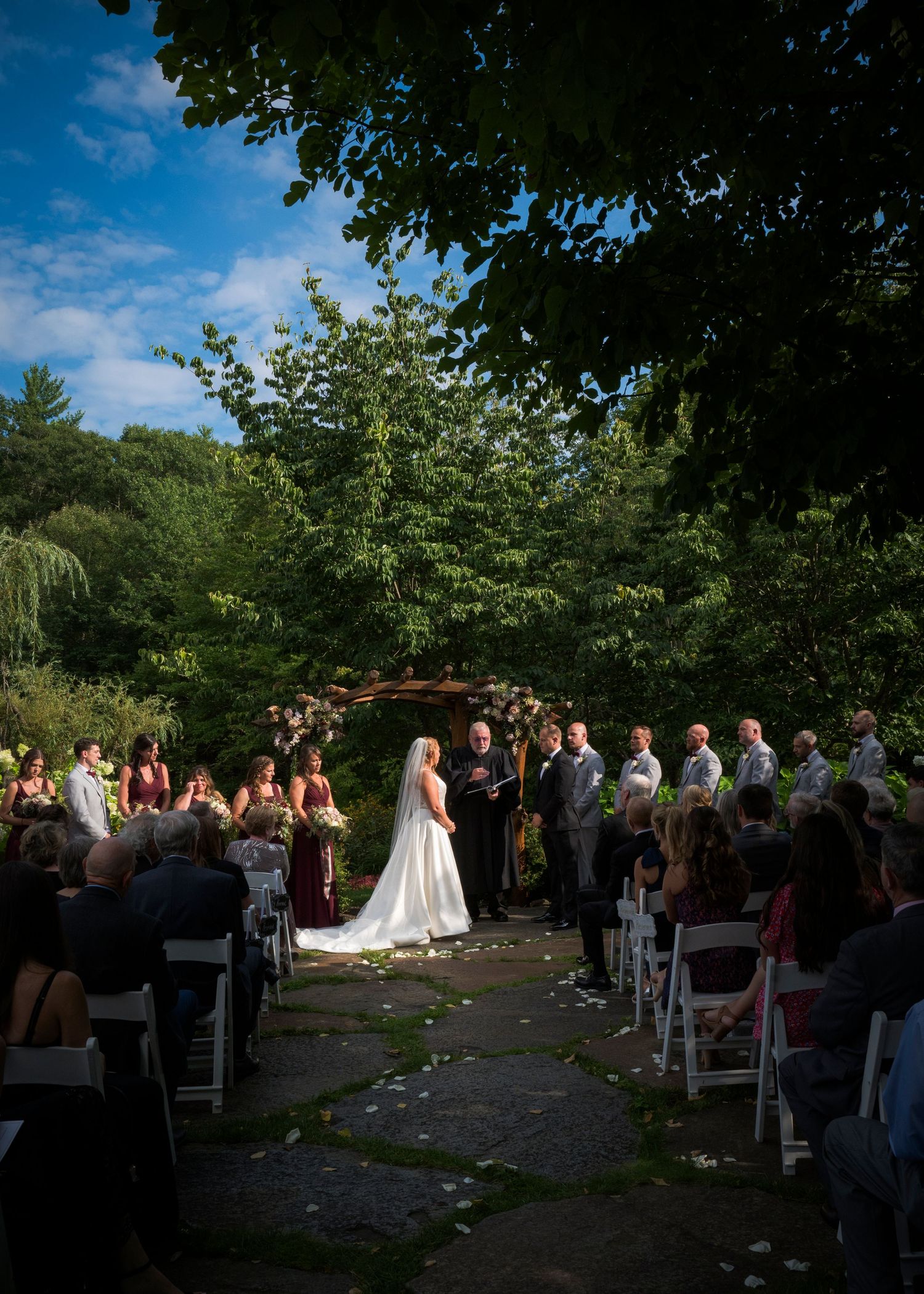 An outdoor wedding ceremony with guests seated on white chairs under green trees.