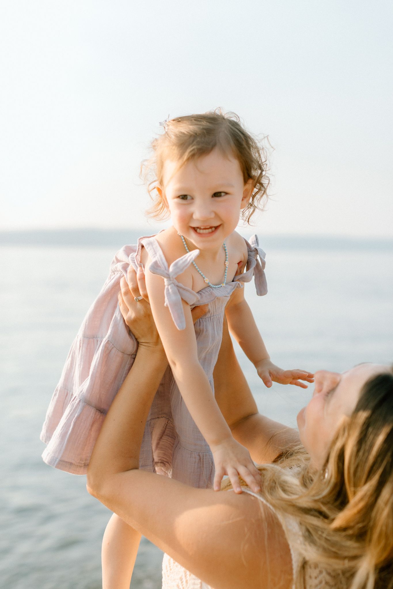 A joyful toddler girl in a pink dress is lifted by her mother on a sunny beach by the water.