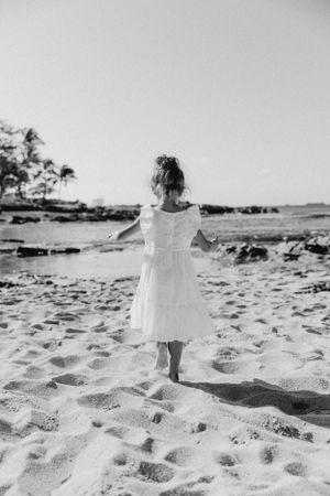 Small child in white dress walking on sandy beach in black and white.
