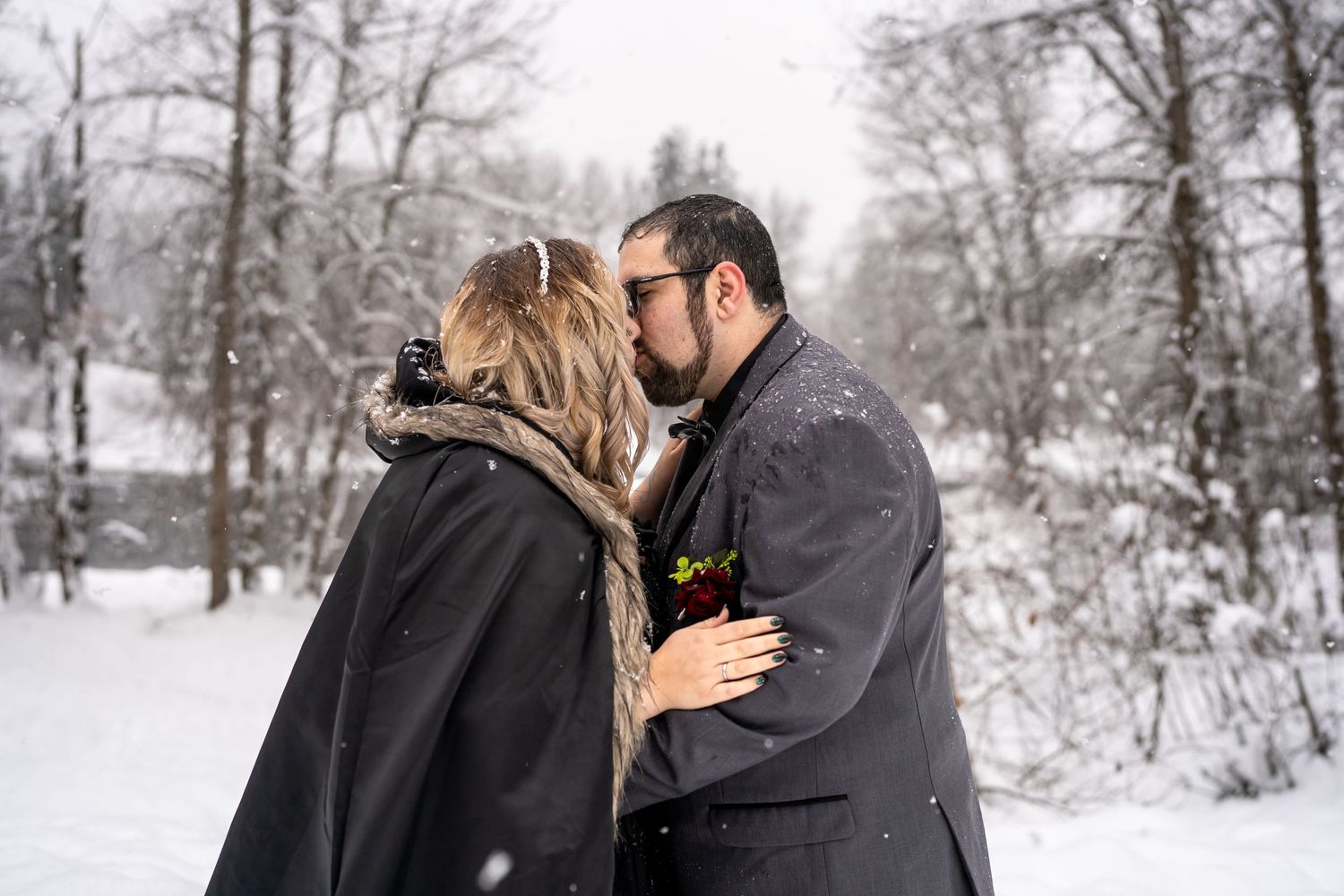Romantic winter moment between couple wrapped in dark coats against snowy forest backdrop.