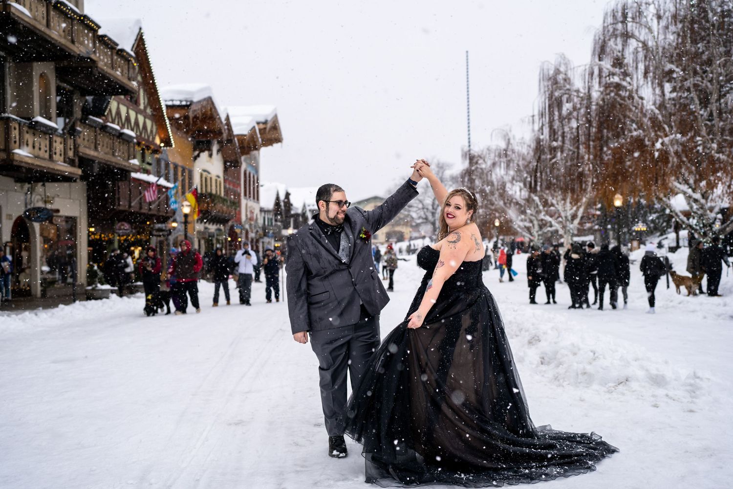 Couple celebrates in the snow along a festive winter street with traditional buildings in background.