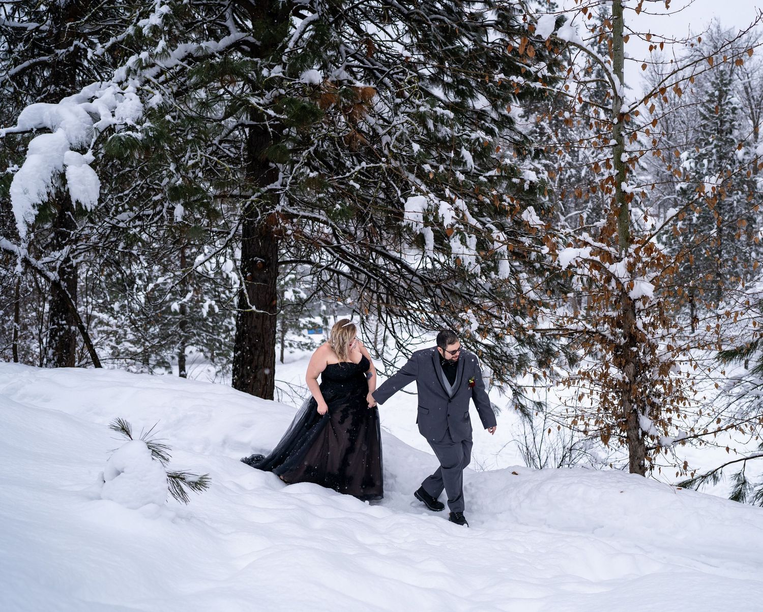 Couple walking through snow-covered forest path wearing formal attire.