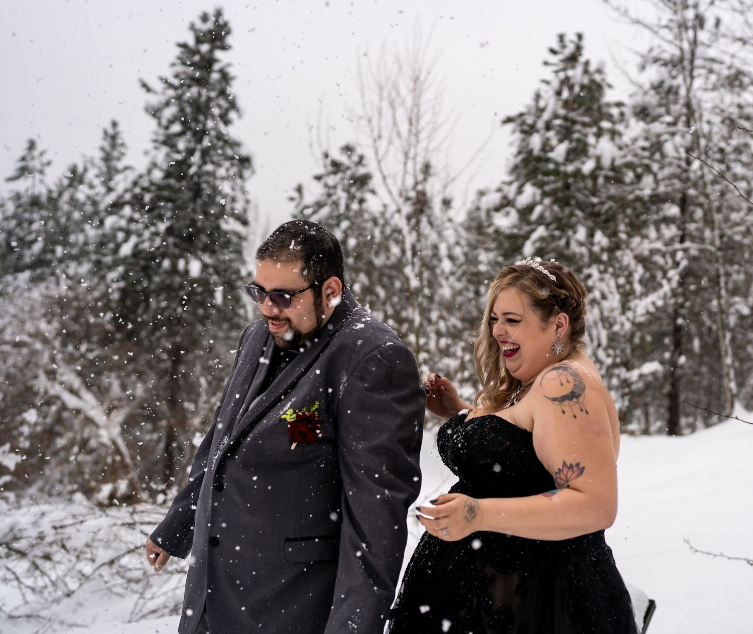 Playful winter photoshoot with couple in formal wear amid snowy pine trees.