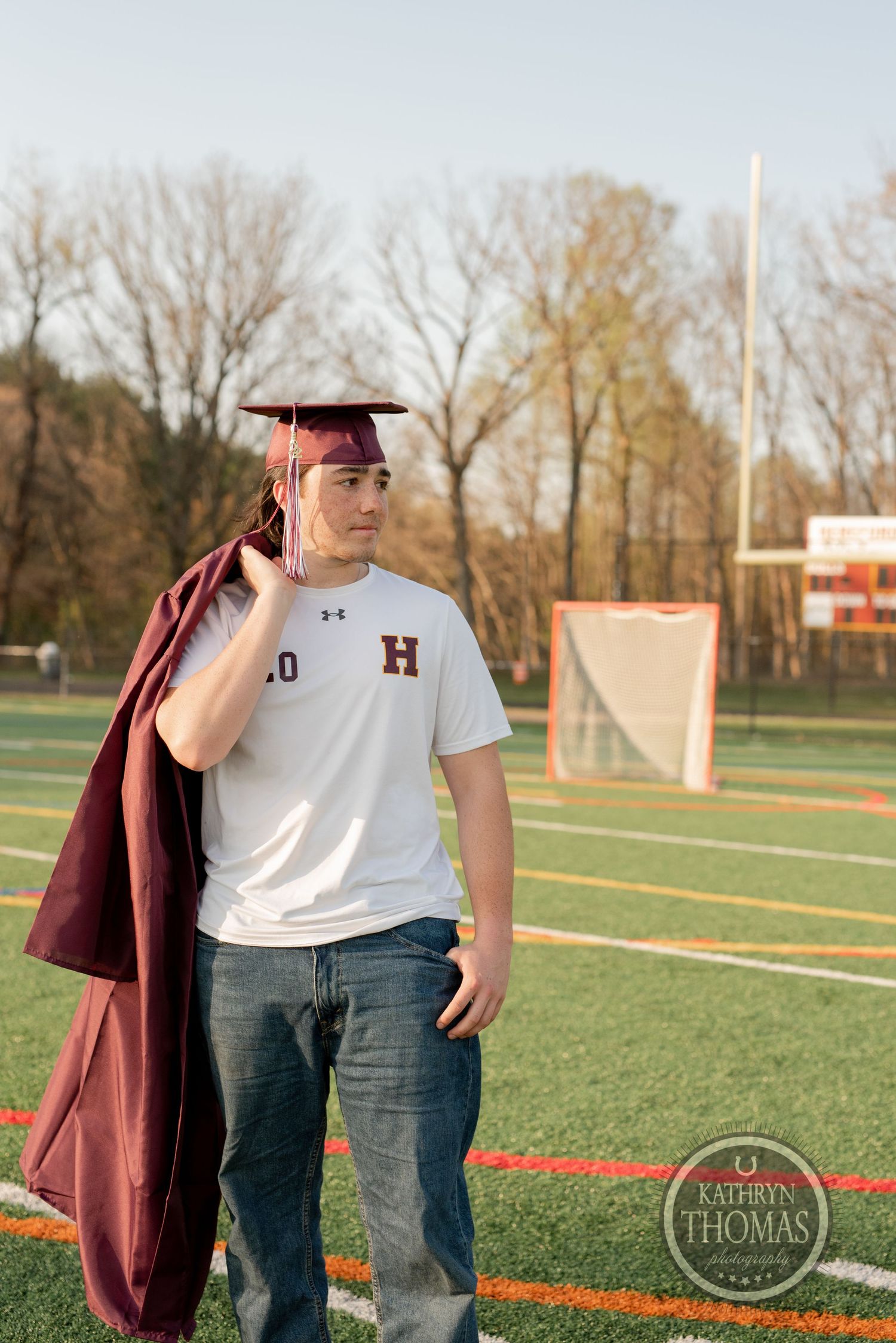Graduate in maroon gown and athletic jersey poses on football field during golden hour photo session.