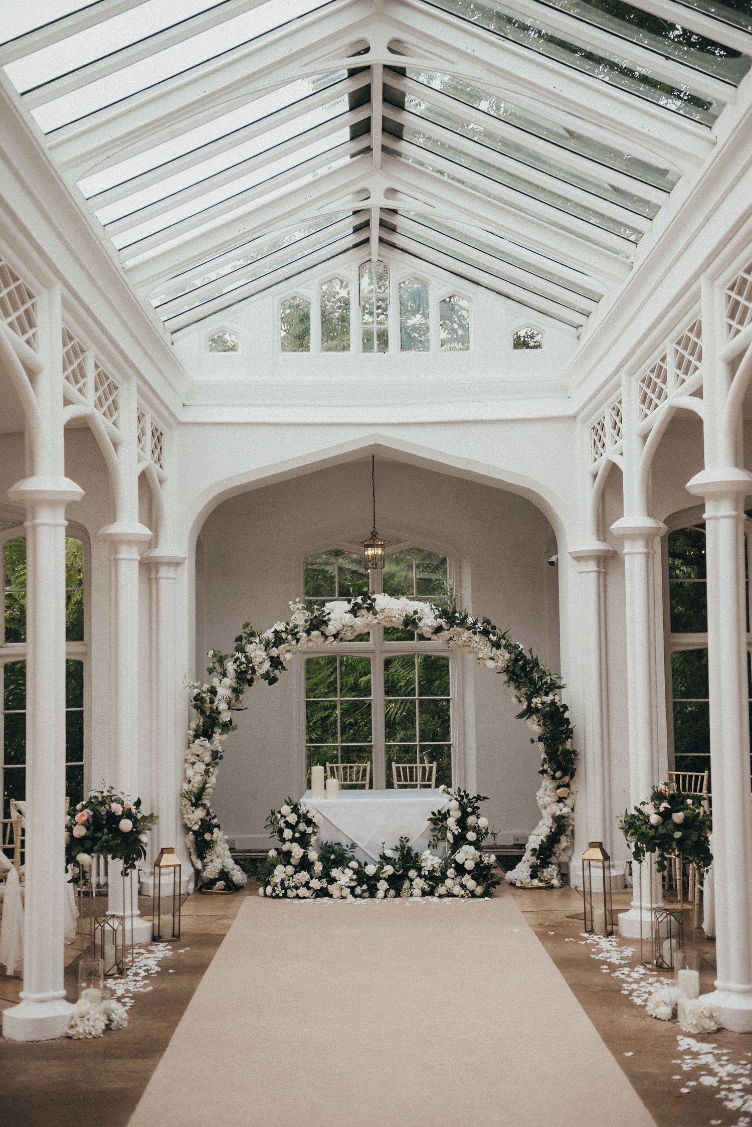 Elegant indoor wedding ceremony setup with a white floral arch and glass ceiling in a conservatory-style venue.